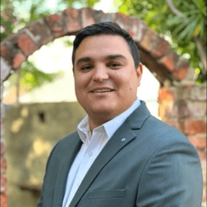 Man in a gray suit jacket and white shirt smiling in front of a rustic brick archway and garden background.