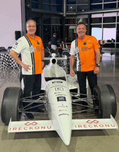 Two men wearing HITEC 2025 racing shirts pose with a branded Formula-style race car indoors at a hospitality tech event.