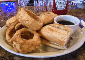 A plate of crispy onion rings and a French dip sandwich with a side of au jus sits on a bar counter, with a ketchup bottle in the background.