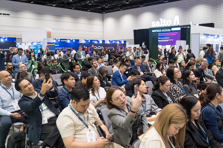 A large audience seated and standing at a busy expo event, many attendees watching a presentation while others take photos or check their phones. Exhibition booths and digital displays are visible in the background.
