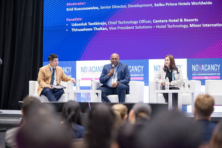 A panel discussion at the NOVACANCY Hotel & Accommodation Industry Expo featuring three speakers seated on stage, with the moderator and discussion topics displayed on a large screen behind them. An audience watches from the foreground.