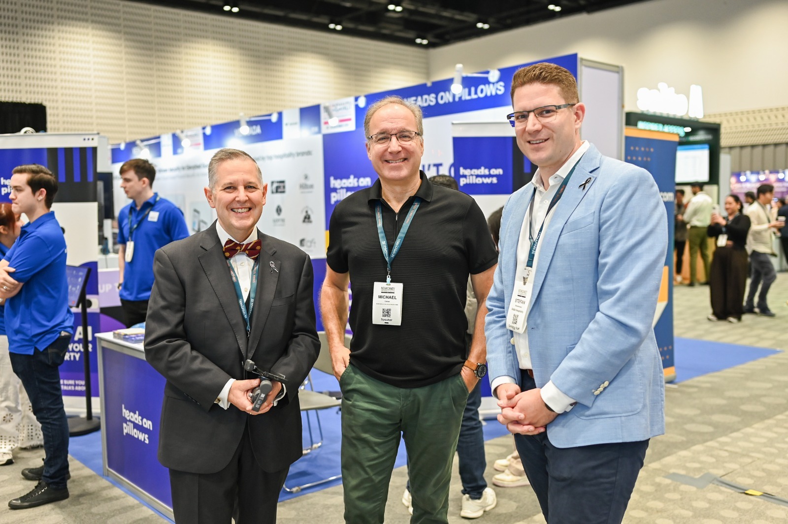 Three men standing and smiling at the camera on an expo floor, with branded booths and attendees visible in the background.